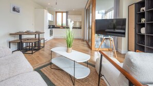 Een moderne woonkamer met een bank, een salontafel en een televisie. Achtergrond is een keuken met een raam en een eettafel.