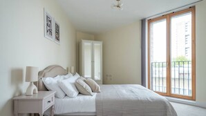 Een slaapkamer met een bed, nachtkastje, lamp en een groot raam met uitzicht op gebouwen.