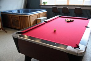Een pooltafel met een rode viltbekleding en een witte poolbal, een houten kast met lades, en een jacuzzi op de achtergrond.