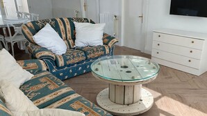 En stue med tapetserede sofaer, et sofabord med glasplade, en hvid kommode og et fjernsyn monteret på væggen.