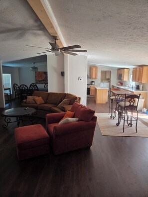 A modern living room with a sectional sofa, a red armchair, a dining area with a table and chairs, and a kitchen with wooden cabinets.
