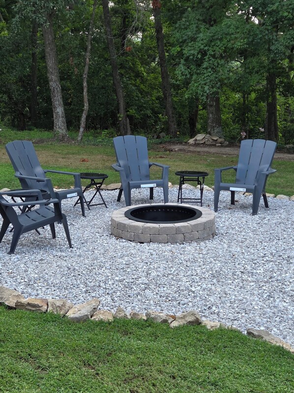 A fire pit surrounded by four Adirondack chairs, set on a gravel area with a grassy border.