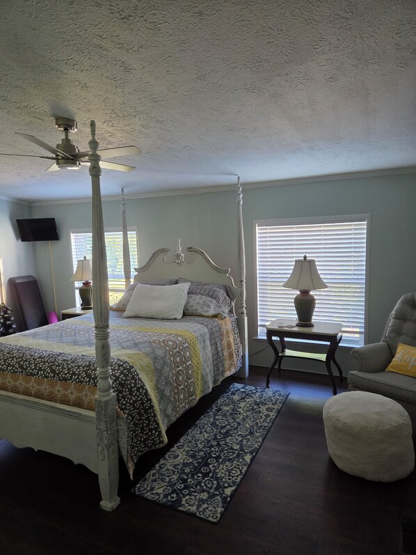 A bedroom with a four-poster bed, a ceiling fan, a television, a window with blinds, a small table with a lamp, and a chair with a cushion.