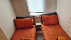 A narrow room with two beds, each with an orange pillow, a small table, and a window with a view. - Cabin Sleeps 4 With Parking (Forfar)