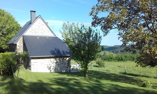 A stone house with a sloped roof, surrounded by lush greenery and a clear sky.