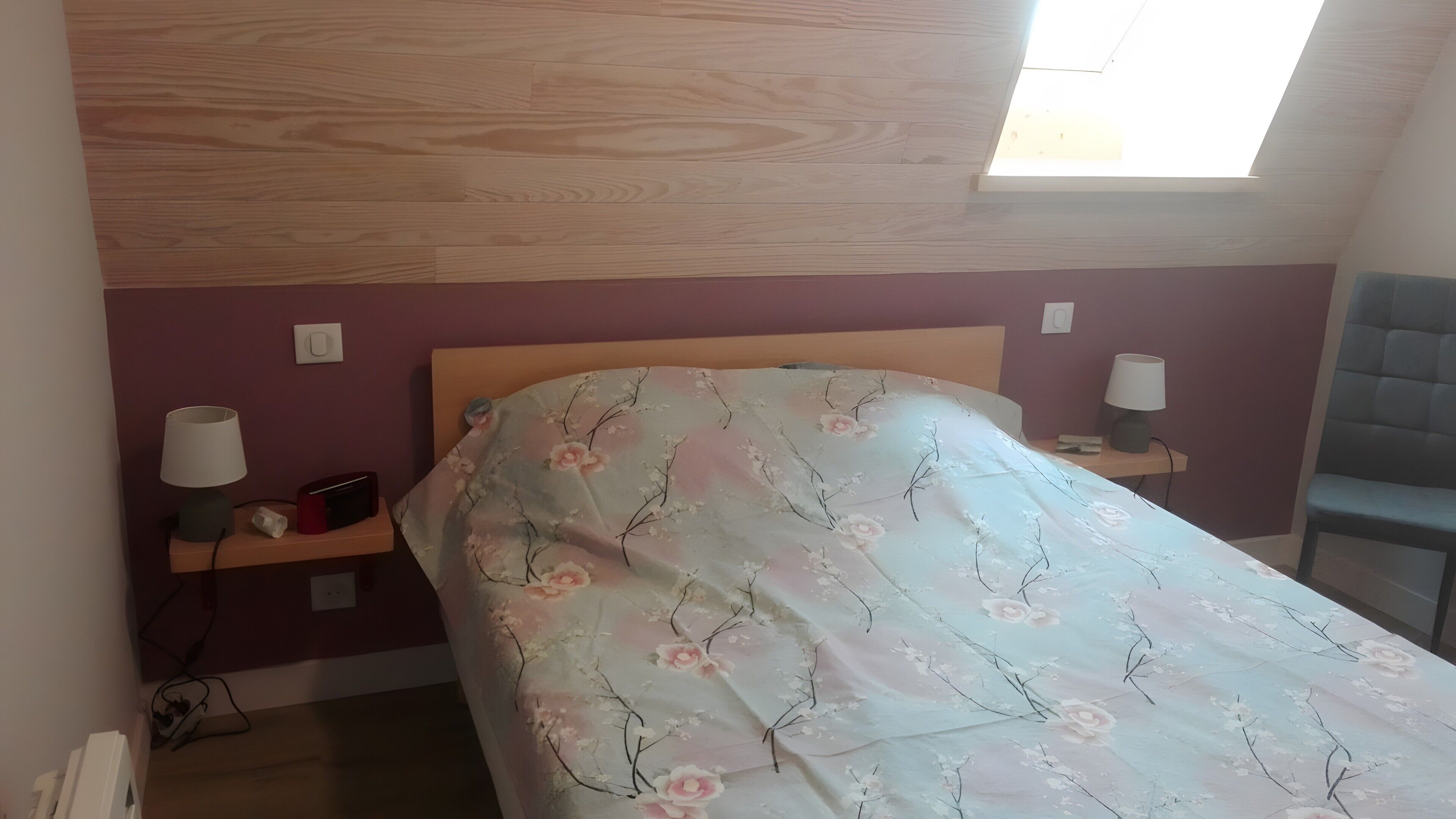 Et soveværelse med træloft, en seng med blomstret sengetæppe, to sengeborde med lamper, og en stol.