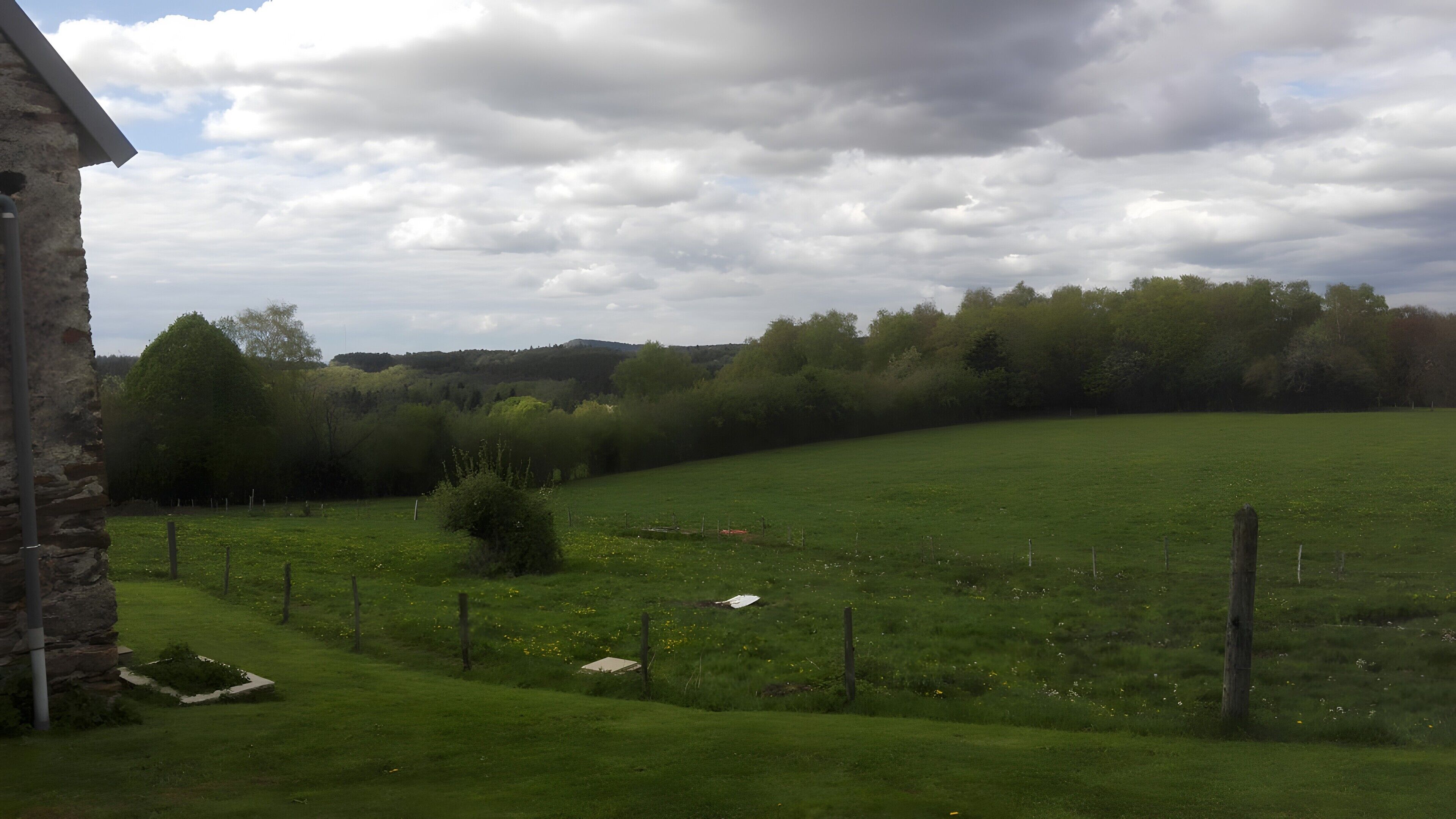 Un paysage rural avec un bâtiment en pierre, un champ clôturé et une rangée d’arbres.