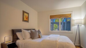 Un dormitorio con cama, mesita de noche, lámpara, ventana con persianas y un cuadro en la pared.