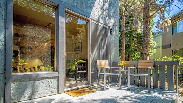 Una terraza de madera con mobiliario exterior, una puerta de cristal y vistas a árboles y un edificio al fondo.
