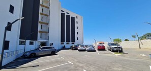 Een parkeerplaats met verschillende auto's, een gebouw met balkons, en een helderblauwe lucht.