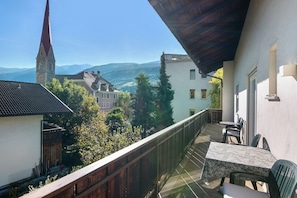 Ein Balkon mit Tisch und Stühlen, mit Blick auf eine Stadt, einen Kirchturm und ferne Berge.