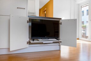 Ein modernes Wohnzimmer mit einem Flachbildfernseher auf einer weißen Unterhaltungsecke, Holzboden und Blick auf einen Balkon.