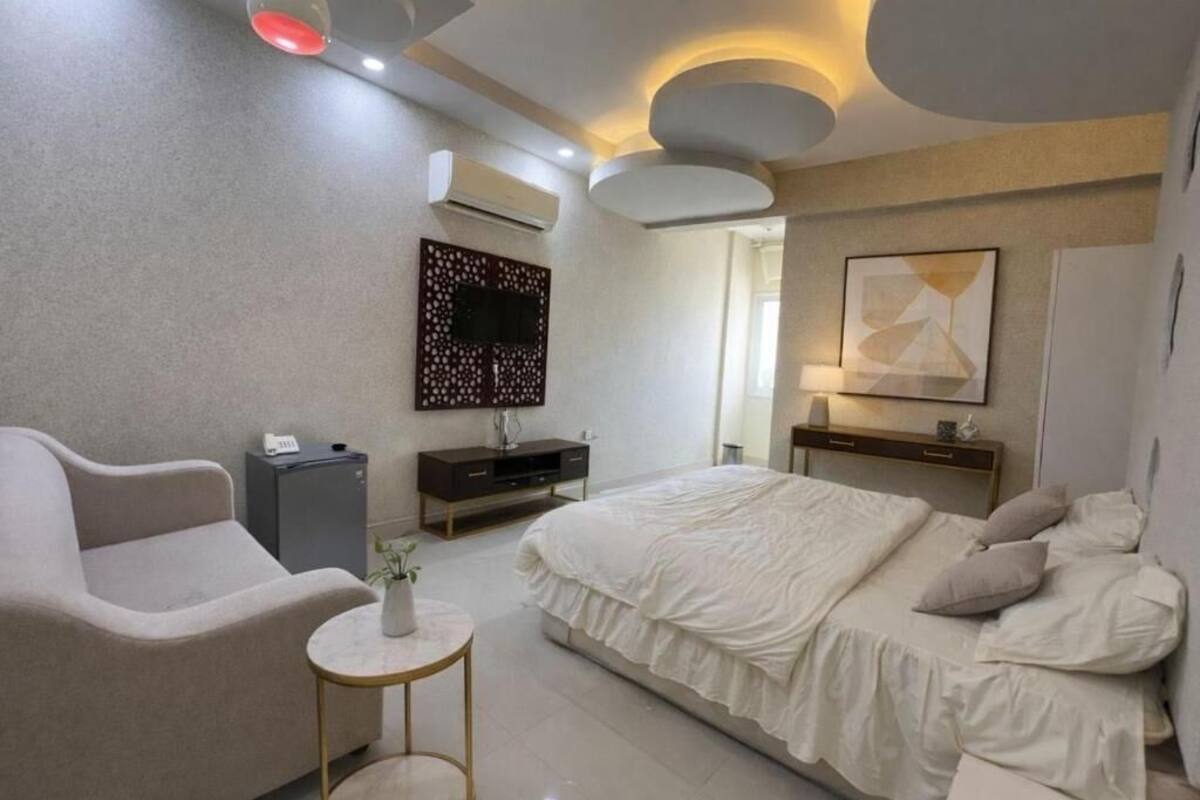 A modern bedroom with a bed, a sofa, a small table, a TV, and a decorative wall piece.