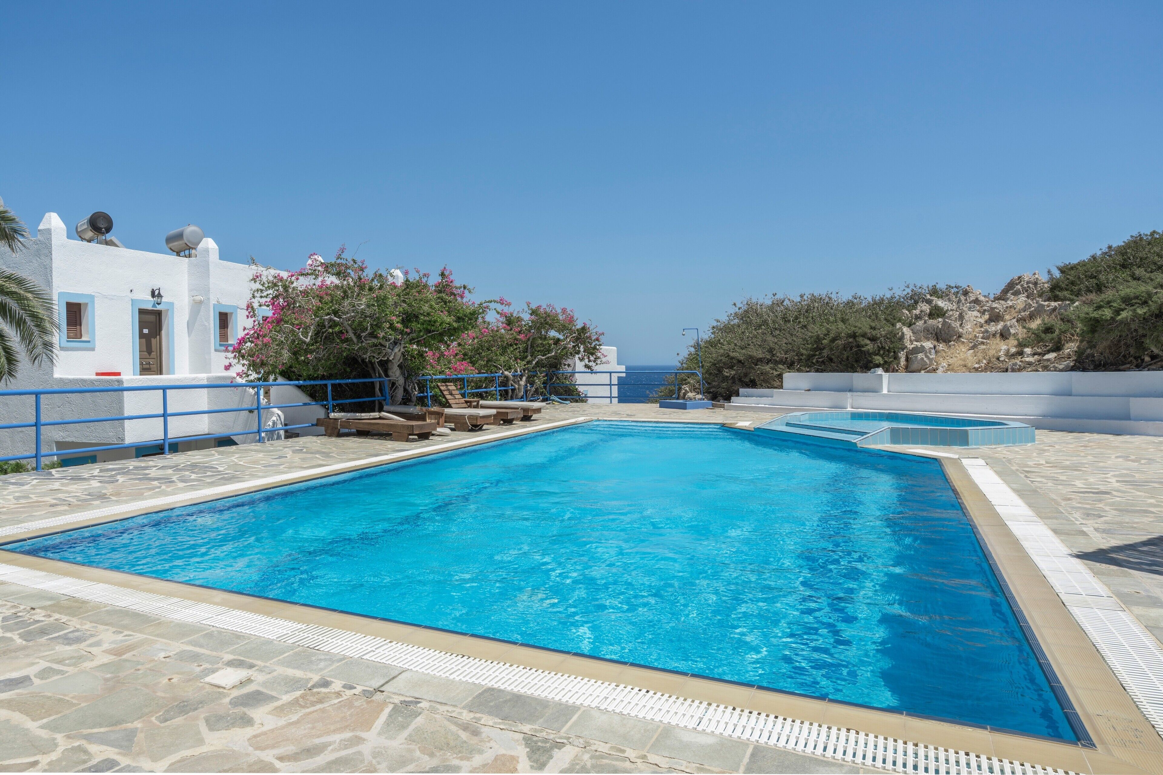 Ein klares, blaues Schwimmbecken mit Steinböden, umgeben von einem weißen Gebäude mit blauen Akzenten und Blick auf das Meer.