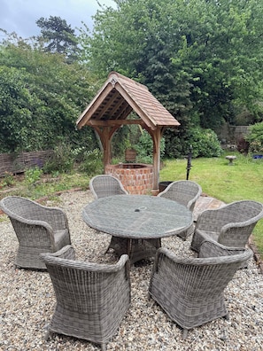 A garden with a wooden gazebo, a round table, wicker chairs, and a brick well.