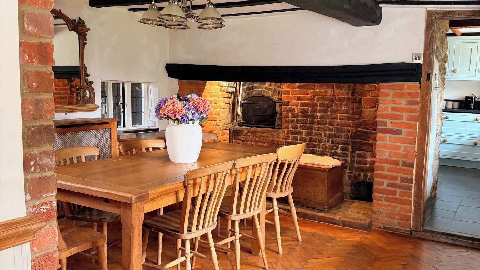 A wooden dining table with chairs, a vase of flowers, and a brick fireplace.