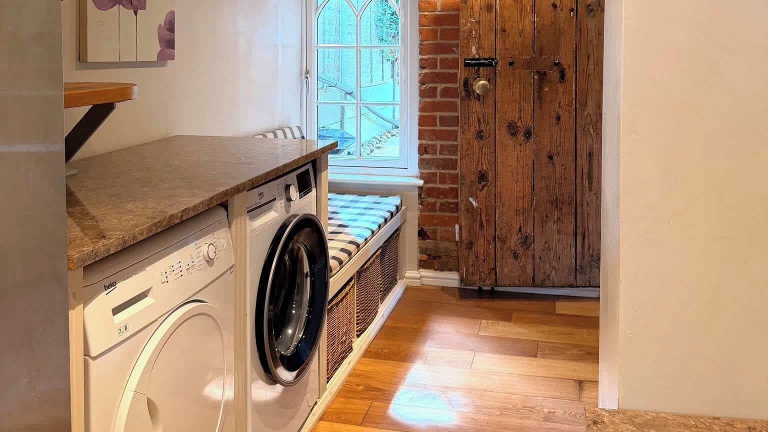 A laundry area with a washing machine, a bench with a striped cushion, a wooden door, and a window with a view of the outside.