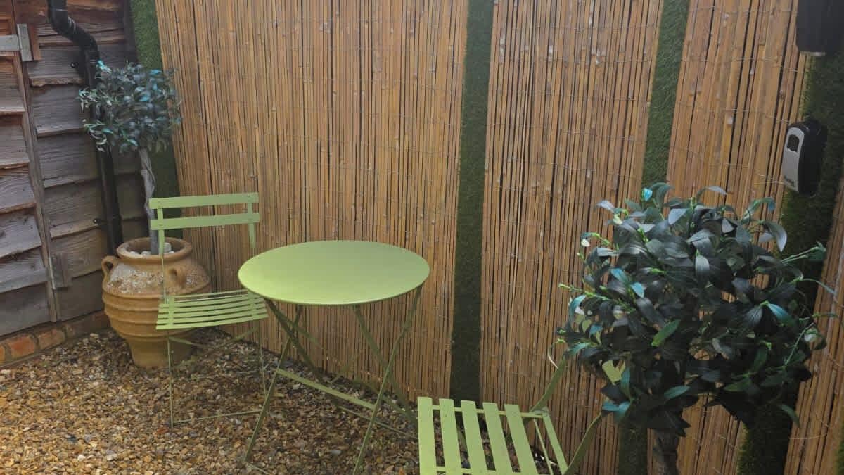 Ein kleiner Sitzbereich im Freien mit einem runden Tisch und zwei Stühlen, umgeben von einem Bambuszaun und Topfpflanzen.