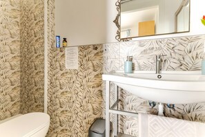 Un baño con papel tapiz estampado con hojas, un lavabo blanco con dos dispensadores de jabón, un espejo y un inodoro.