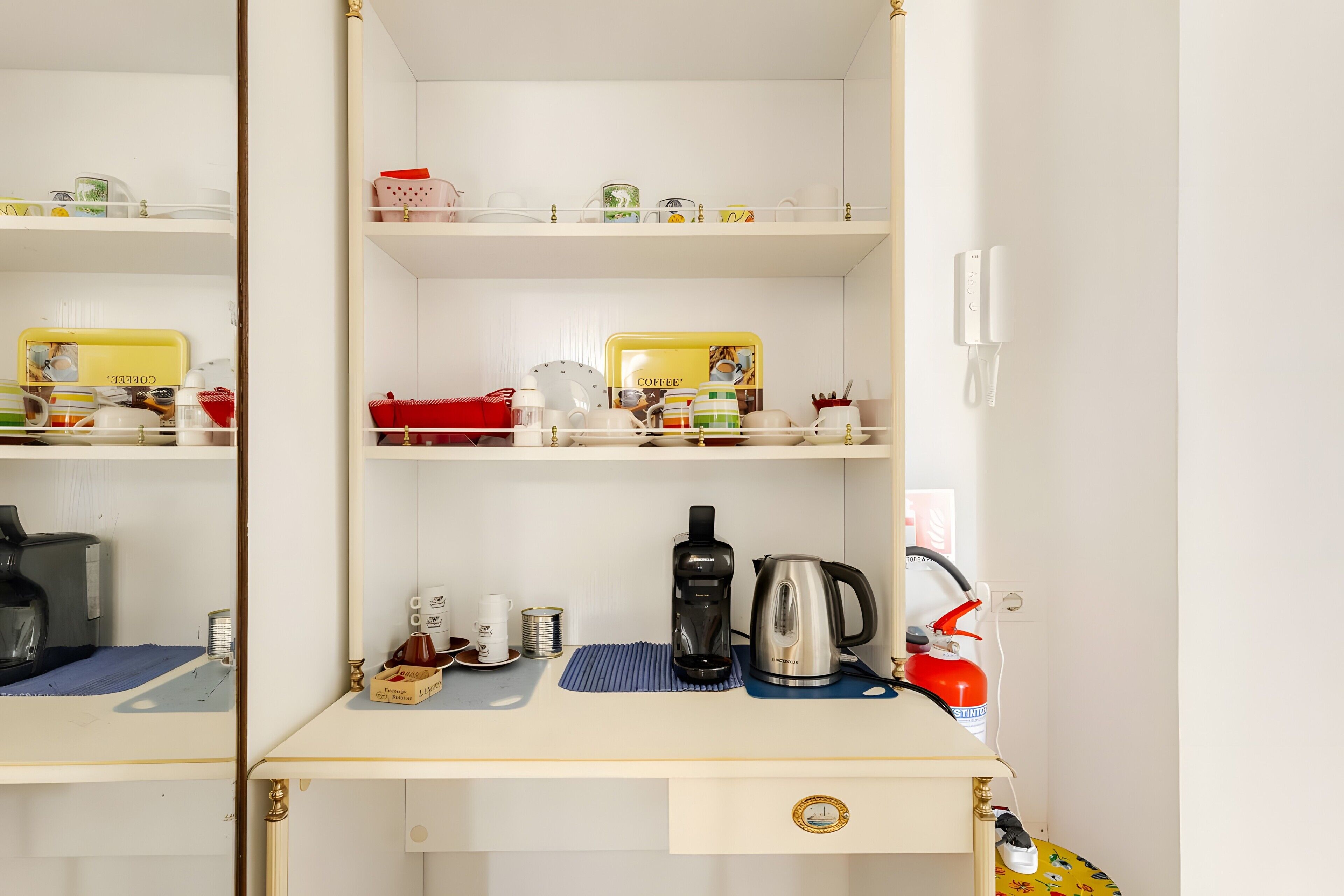 Een hotelkamer met een wit bureau, een koffiezetapparaat, een waterkoker en een rode brandblusser.