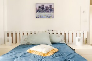 Un lit recouvert d’une couvre-lit bleue et orné d’un oreiller blanc à volants jaunes, une table de chevet blanche avec une lampe blanche, et un tableau représentant des bâtiments, accroché au mur.
