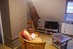 Uma sala de estar com televisão, uma mesa de madeira com um tabuleiro de xadrez, um sofá com almofadas estampadas, uma cadeira de madeira e um guarda-roupa.