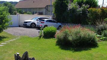 Zona residencial con un coche blanco estacionado en una entrada, una casa con techo de tejas y un césped con arbustos florecientes.