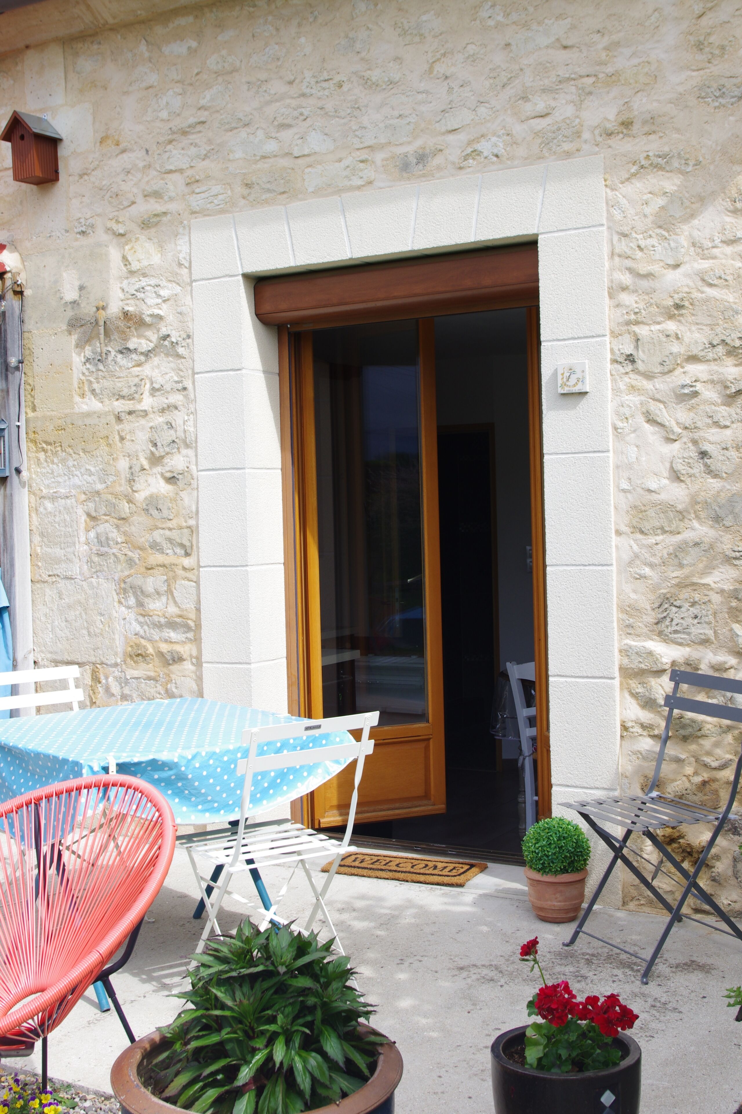 A stone building with a wooden door, a blue table with a polka dot tablecloth, a red chair, and potted plants.