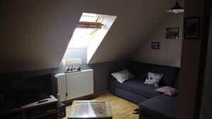 Ein Zimmer mit schrägem Dach, einem Fernseher, einem Glastisch, einer Couch und einer Heizung.
