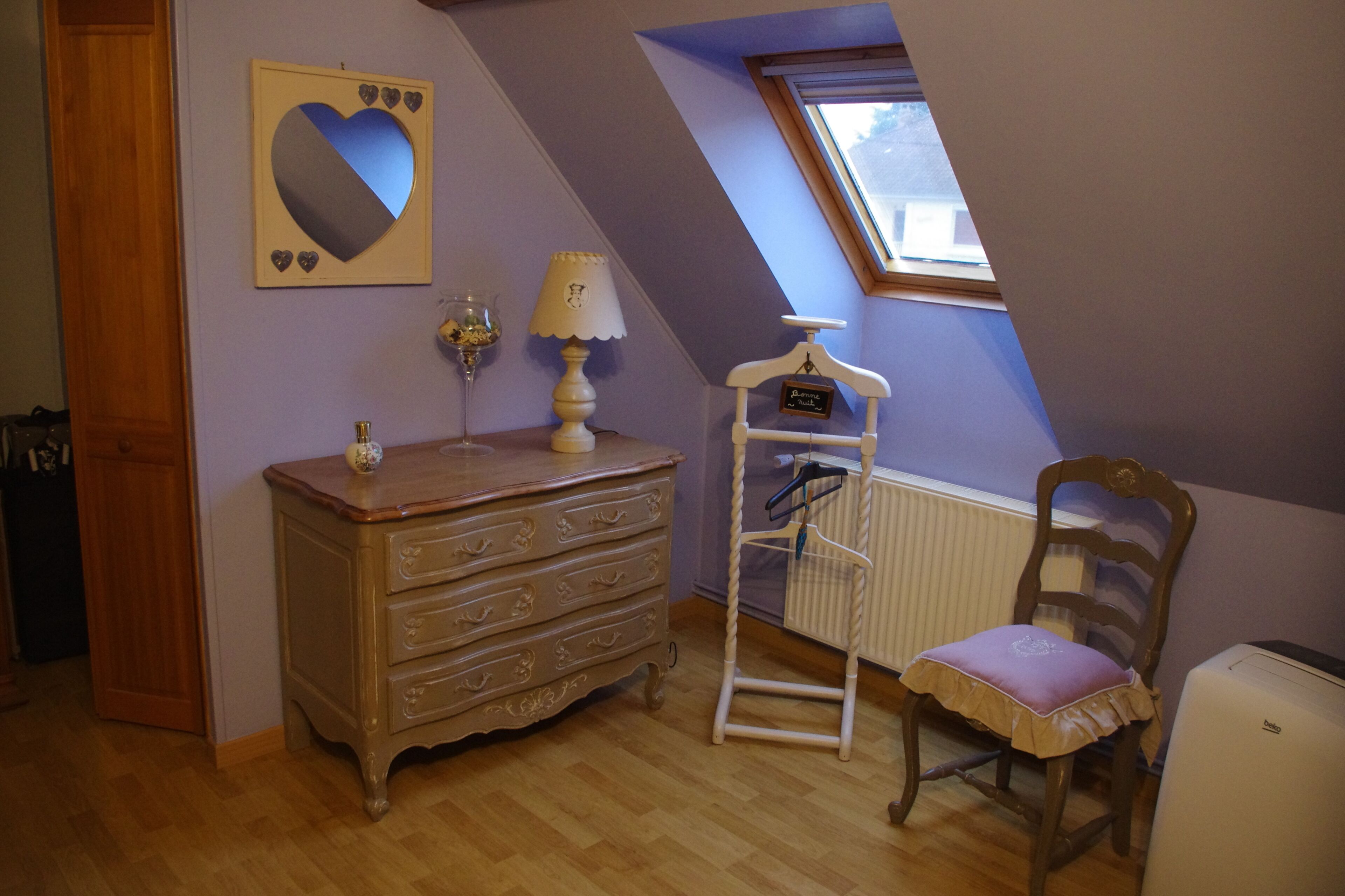 Une pièce comprenant une commode en bois, une chaise avec un coussin, une lampe, un miroir en forme de cœur et une fenêtre.