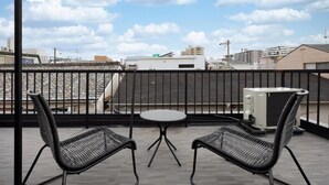 A balcony with two chairs and a small table, overlooking a cityscape with buildings and a clear sky. - JAPAVISTA Hinata (KONOHANA WARD)
