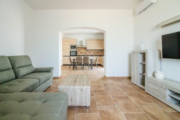 Ein Wohnzimmer mit einer Couch, einem Couchtisch und einem Fernseher auf einem Schrank. Durch einen Bogen ist eine Küche mit Schränken und einem Essbereich erkennbar.