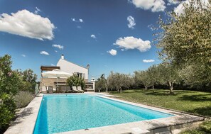 Blick vom Pool auf ein Haus mit Schwimmbad, umgeben von viel Grün und blauem Himmel.