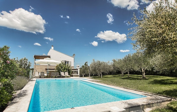 Blick vom Pool auf ein Haus mit Schwimmbad, umgeben von viel Grün und blauem Himmel.