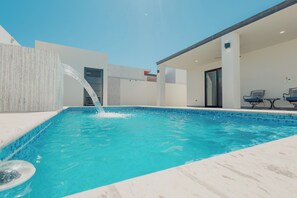 Ein Schwimmbecken mit Wasserspiel, ein überdachter Bereich mit Sitzgelegenheiten und ein modernes architektonisches Design.