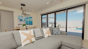 A modern living room with a grey sectional sofa, starfish pillows, a dining area, and a large window with a pool view. - Casa Luna  By FMI Rentals (Puerto Peñasco)