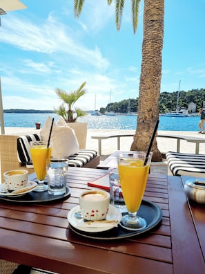 Ein Tisch mit Getränken und einer Tasse, mit Blick auf ein Gewässer mit Booten und einem Baum.