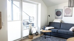 Un salon moderne avec un canapé gris, une petite table en bois et un tapis gris. Une grande fenêtre offre une vue sur le mobilier d’extérieur.