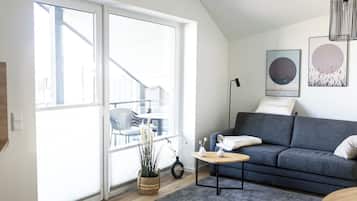 Ein modernes Wohnzimmer mit einem grauen Sofa, einem kleinen Holztisch und einem grauen Teppich. Ein großes Fenster bietet Blick auf Gartenmöbel.