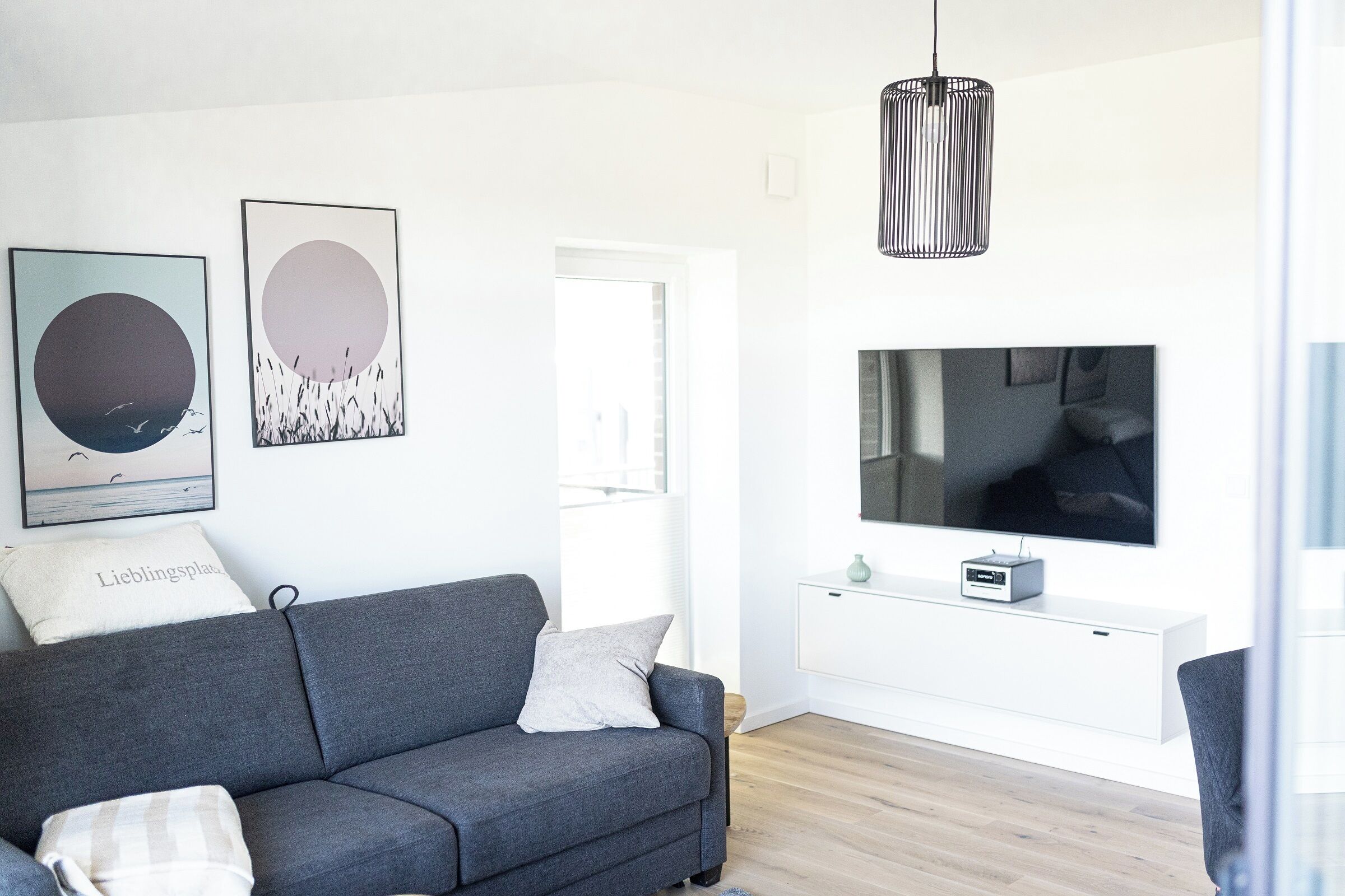 Ein modernes Wohnzimmer mit grauem Sofa, zwei Wandbildern, einer schwarzen Pendelleuchte, einem Flachbildfernseher und einem kleinen weißen Schrank.