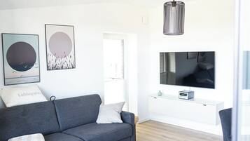 Ein modernes Wohnzimmer mit grauem Sofa, zwei Wandbildern, einer schwarzen Pendelleuchte, einem Flachbildfernseher und einem kleinen weißen Schrank.