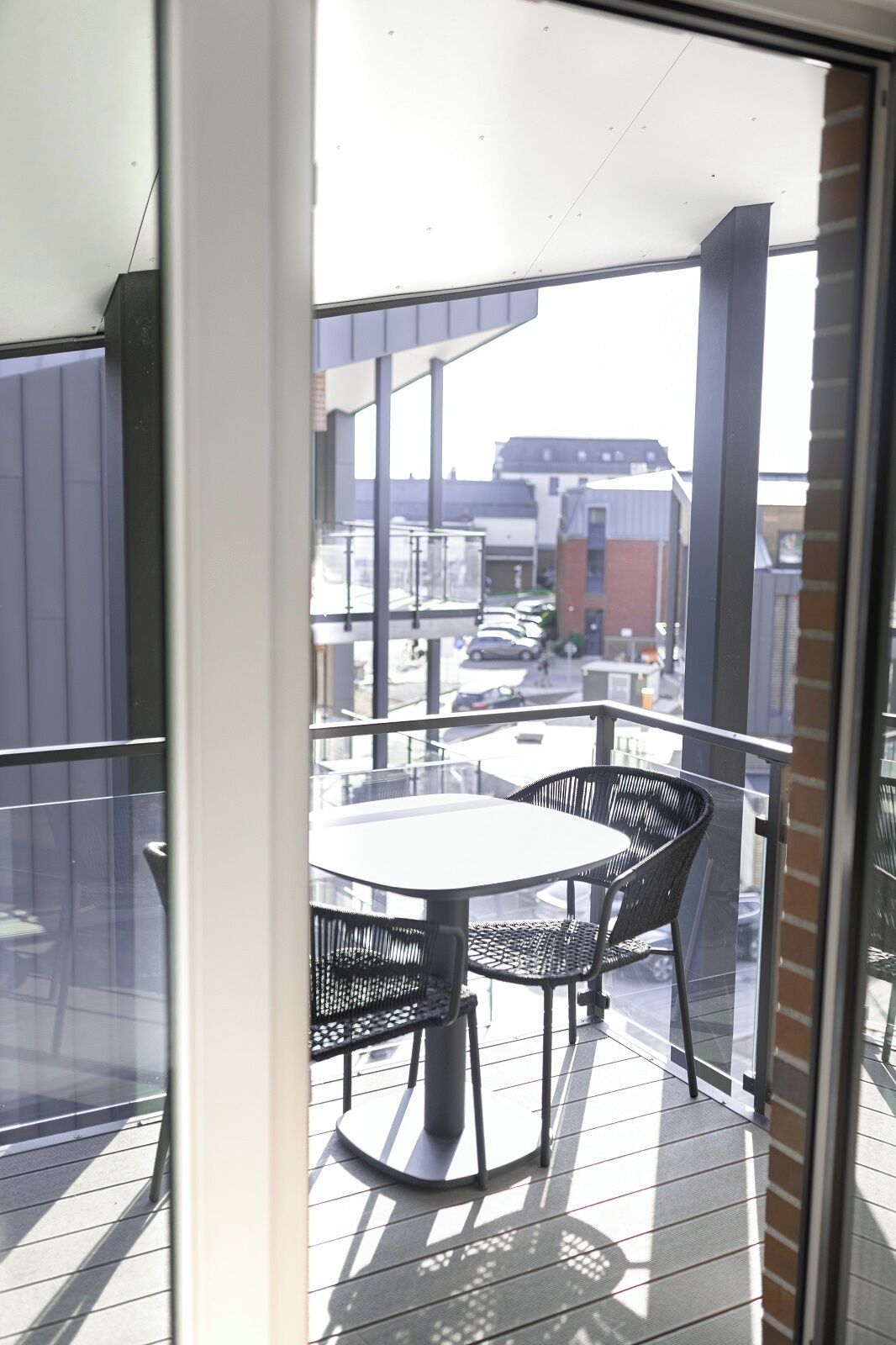 Un balcon avec une table et des chaises, donnant sur une rue bordée de voitures garées et d’immeubles.
