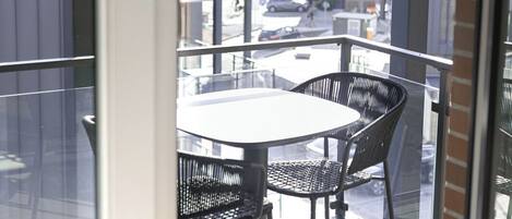Un balcon avec une table et des chaises, donnant sur une rue bordée de voitures garées et d’immeubles.