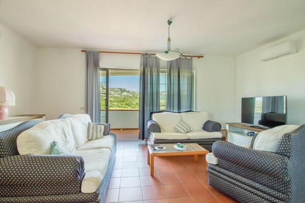 A living room with a sofa, a patterned armchair, a coffee table, a television, and a view of the outdoors. - GuestHost - Cozy Apartment by the Vineyard (Arzachena)