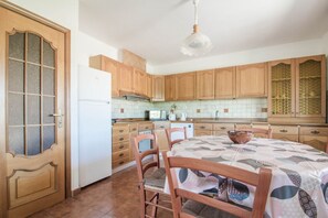 A kitchen with wooden cabinets, a white refrigerator, a dining table with chairs, and a hanging light fixture. - GuestHost - Cozy Apartment by the Vineyard (Arzachena)