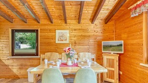 A wooden cabin interior with a dining table set for two, a television on a cabinet, and a window with a view of greenery.