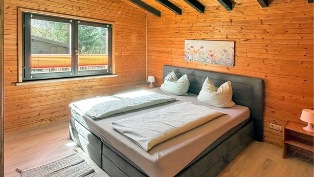 A wooden cabin bedroom with a bed, pillows, a nightstand, a lamp, and a window with blinds.