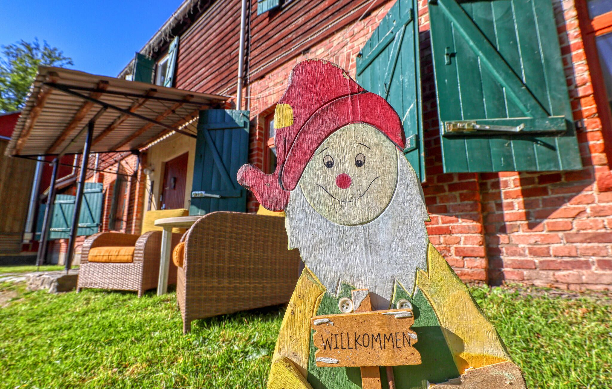Un gnome en bois, avec une pancarte sur laquelle on peut lire « WILLKOMMEN », se trouve devant un bâtiment en briques doté de volets verts.