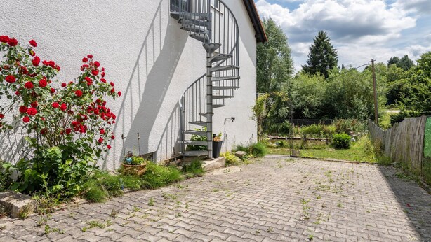 En hvid bygning med en spiraltrappe, der fører til et vindue, omgivet af grøn vegetation og en belagt sti.