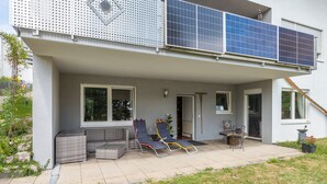 Et hus med en overdækket terrasse, to liggestole, et lille bord og solpaneler på taget.